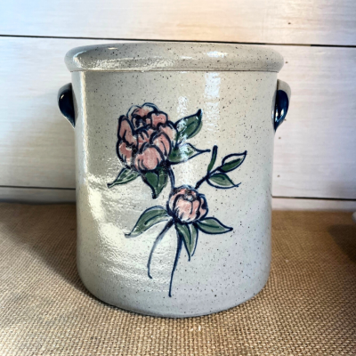                             wheel thrown 1 gallon crock with a spring peony pattern handpainted in blush pink with cobalt lining and greenery on a white shiplap background

