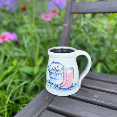                     hand thrown stoneware mug pictured in a garden on a wooden chair.  The mug is hand decorated in cobalt blue with red garden boots and a harvest basket of fresh picked apples        