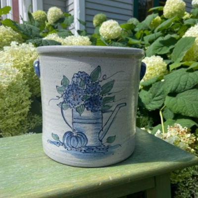                             1 gallon hand thrown stoneware crock hand painted with a watering can, hydrangeas and pumpkin in a classic cobalt blue, photo'd in a garden of hydranges.